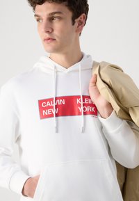 Hanorac alb din bumbac, cu un grafic dreptunghiular roșu având textul "CALVIN NEW YORK", cu sfori de ajustare și o buzunar frontal.