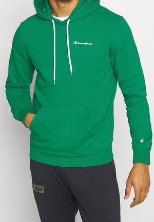 Man wearing green Champion hoodie with white drawstrings and logo, black pants with patch on thigh, hand in hoodie pocket.