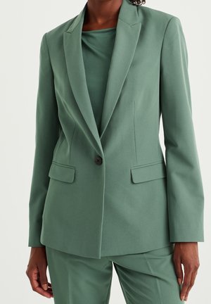 Frau in einem maßgeschneiderten salbeigrünen Blazer mit passenden Hosen und einem Rundhalstop, stehend vor einem schlichten, hellen Hintergrund.