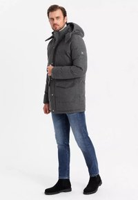 Giacca imbottita grigia con cappuccio, bottoni frontali e due tasche a bavero. Abbinata a jeans blu e stivaletti neri, su uno sfondo bianco semplice.