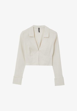 Camicetta a maniche lunghe tagliata in tessuto beige chiaro. Presenta un colletto classico, scollatura a V profonda e chiusura frontale con bottoni. Texture liscia.