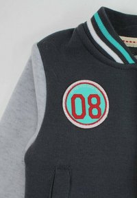 Veste universitaire gris foncé et gris clair avec col rayé et écusson rond présentant "08" en rouge sur fond turquoise.