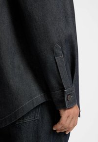 Manica di camicia in denim scuro con polsino abbottonato e una mano che si posa vicino alla tasca di jeans in denim scuro.