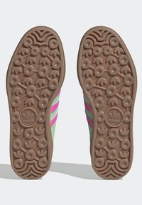 Vihreät ja vaaleanpunaiset Adidas-tennarit, joissa on teksturoitu ruskea kumipohja, jossa on pyöreitä kuvioita, sekä kolme paksua vaaleanpunaista raitaa sivuilla.