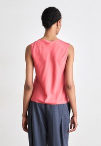 Κοράλι σατέν αμάνικο τοπ με απαλό υλικό και στρογγυλή λαιμόκοψη, συνδυασμένο με χαλαρούς γκρίζους παντελόνες, προβαλλόμενο από πίσω.