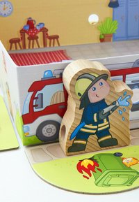Holzfigur eines Feuerwehrmanns, die einen lächelnden Charakter in einer blauen Uniform mit gelben Streifen darstellt, der einen Schlauch hält und in der Nähe einer Abbildung eines Feuerwehrautos positioniert ist.
