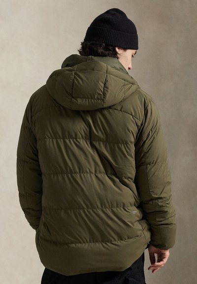 Casaco verde acolchoado com design almofadado e capuz ajustável. Usado com um gorro de malha preto. Apresenta costuras horizontais e um corte confortável.