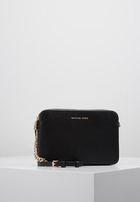 Černá crossbody taška Michael Kors se zlatým řetízkem a přezkou na bílém podstavci na jednoduchém šedém pozadí.