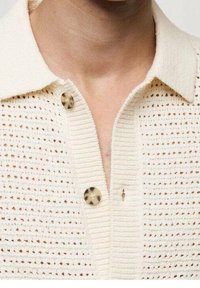 Polo bianco in maglia con un motivo testurizzato, dotato di colletto, due bottoni rotondi beige e un piccolo bottoncino anteriore.
