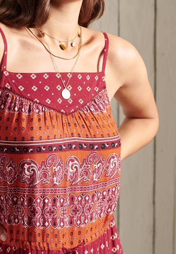 Femme portant une robe rouge-orange sans manches à motifs et des colliers en or superposés, debout une main sur la hanche.