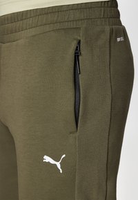 Olivgrüne Jogginghose aus weichem Stoff, mit einer schwarzen Reißverschlusstasche und einem weißen Puma-Logo auf der linken Seite.