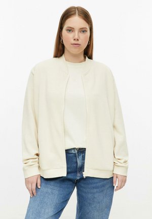 Giovane donna con capelli rossi lisci che indossa una giacca zip-up testurizzata color avorio, camicia bianca e jeans blu, rivolta in avanti su sfondo bianco.