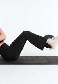 Personne en leggings noirs et baskets blanches fait de l’exercice sur un tapis noir avec une petite balle d’exercice noire tenue entre les mollets pliés.