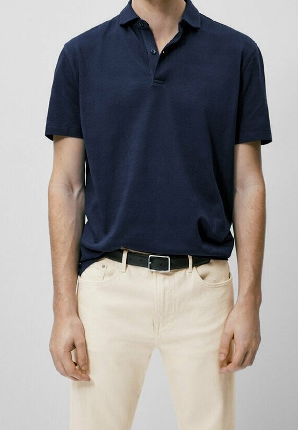Homme portant un polo bleu marine à manches courtes rentré dans un pantalon beige avec une ceinture noire, debout devant un fond uni.