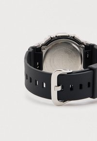 Μαύρο λαστιχένιο λουράκι ρολογιού με υφή στην επιφάνεια, αγκράφα από ανοξείδωτο ατσάλι και εκτεθειμένη μεταλλική πλάτη κάσας που υποδεικνύει αντοχή στο νερό και αντοχή σε κραδασμούς.