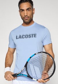 Camiseta de manga corta azul claro con el logo "LACOSTE" en impresión oscura. El modelo sostiene una raqueta de tenis negra con detalles en azul.