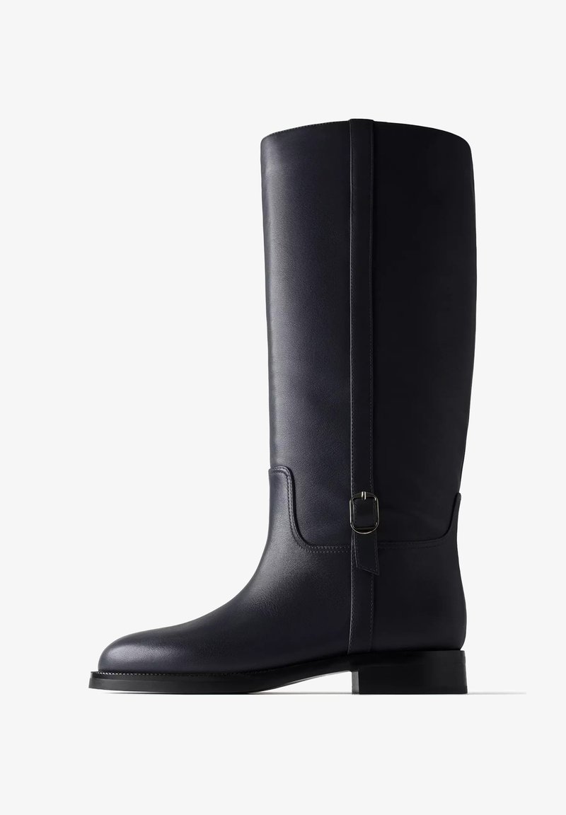 Botte haute en cuir noir avec un talon bas et une sangle décorative avec une petite boucle argentée sur le côté extérieur.