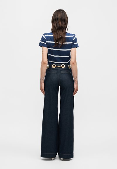 Γυναίκα που κοιτάζει από την πλάτη, φοράει μπλε και λευκό ριγέ πουκάμισο, σκούρα τζιν με φαρδιά μπατζάκια και ζώνη με χρυσή αλυσίδα.