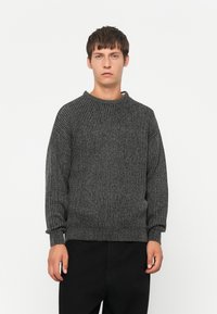 Giovane uomo con capelli corti castani in piedi di fronte, indossa un maglione a coste grigio scuro e pantaloni neri su uno sfondo bianco.