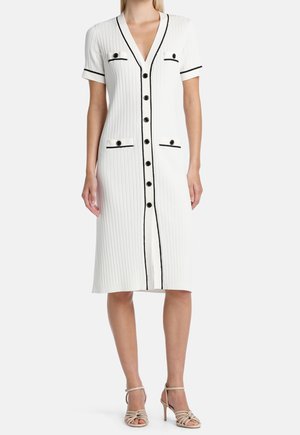 Robe blanche côtelée longueur genoux, manches courtes, détails de boutons noirs, bordure noire sur les poches et talons ouverts beiges à brides à la cheville.