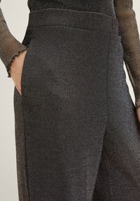 Pantalon noir avec un subtil éclat métallique, présentant une texture lisse, des poches latérales et une coupe droite. Détails de taille visibles.