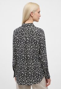 Zwart-wit gebloemde blouse met lange mouwen en een kraag, gemaakt van een luchtige stof met een plooi aan de achterkant. De zoom is licht gebogen.