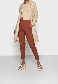 Beige trenchcoat over een beige geribbeld sweater, gecombineerd met roestkleurige broeken met een gestrikte taille, en lichtgekleurde sandalen met banden.