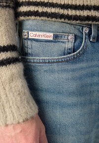 Calvin Klein traperice u svijetloplavoj boji s klasičnim krojem. Ima oznaku od tkanine i bočnu džep s srebrnim detaljima. Rukav vune je vidljiv.