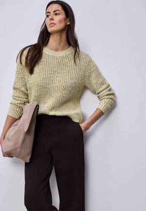 Žena s dlouhými tmavými vlasy, která nosí žluto-zelený pletený svetr a černé kalhoty, drží béžovou tašku přes rameno a jednou rukou má ruku v kapse.