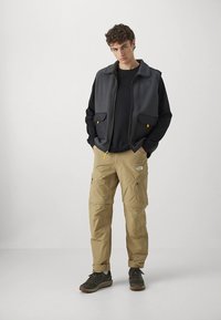 Grijs-zwarte bodywarmer met rits over een zwart shirt, beige cargobroek en donkere schoenen; heeft zakken en een logo op de bodywarmer.