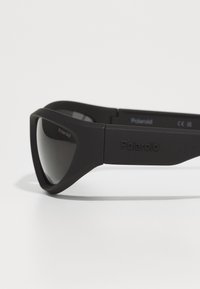 Mustat Polaroid-aurinkolasit, jossa on matta pinta, kulmikas muotoilu ja tummat linssit; sisältää brändäyksen varressa ja saranassa.