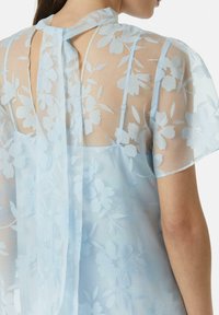 Jasnoniebieska, przezroczysta bluzka z kwiatowym wzorem, półprzezroczysty materiał, krótkie rękawy oraz dekolt w kształcie dziurki z wiązaniem.