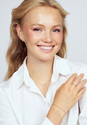 Ung kvinna med blont hår ler, iklädd en vit skjorta och delikat guldsmide inklusive ett halsband, armband, örhängen och ring.