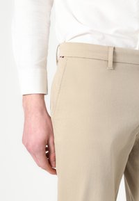 Beige gemusterte Hose mit einer subtilen Struktur, ausgestattet mit Seitentaschen und Gürtelschlaufen, kombiniert mit einem weißen Langarmhemd.