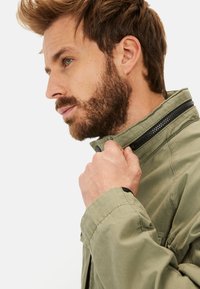 Olive green jacket with a high collar, zip closure, and subtle black accents. Made of textured fabric, featuring a relaxed fit.