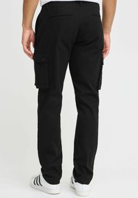 Pantalons cargo noirs avec poches latérales, fermetures à bouton et coupe décontractée. Portés avec des baskets blanches à rayures noires.