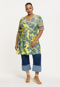 Gelbe Tunika mit blauen Paisley- und Blumenmustern, V-Ausschnitt, kurze Ärmel; kombiniert mit knöchelfreien Jeanshosen und gelben Riemchensandalen.