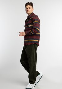 Camisa de botones de rayas rojas y marinas con dos bolsillos en el pecho; combinada con pantalones de pana verde oscuro y zapatillas blancas.