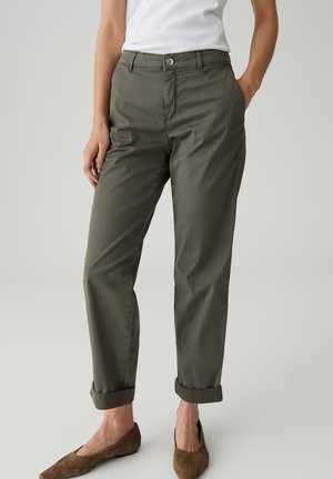 Frau trägt hochgekrempelte olivgrüne Chinos, weißes ärmelloses Oberteil und braune Wildlederflats, steht mit einer Hand in der Tasche.