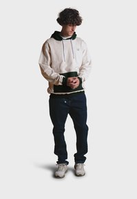 Sudadera beige con detalles en verde oscuro, cordones y un bolsillo frontal. Combinada con jeans azul oscuro y zapatillas deportivas grises. Estilo de moda urbana casual.