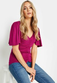 Blusa fucsia con mangas cortas abullonadas, escote en forma de V y detalle fruncido en la parte delantera. Combinada con jeans azules, sentada en un taburete.