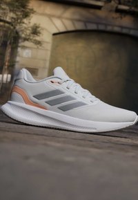 Chaussure de sport blanche avec des rayures grises et des accents pêche reposant sur une surface en pierre à l'extérieur, montrant le profil latéral et les lacets.