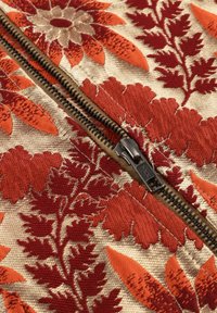 Geborduurd stof met grote rode en oranje bloemmotieven op een beige achtergrond, met een zichtbare zwarte metalen ritsdetail.