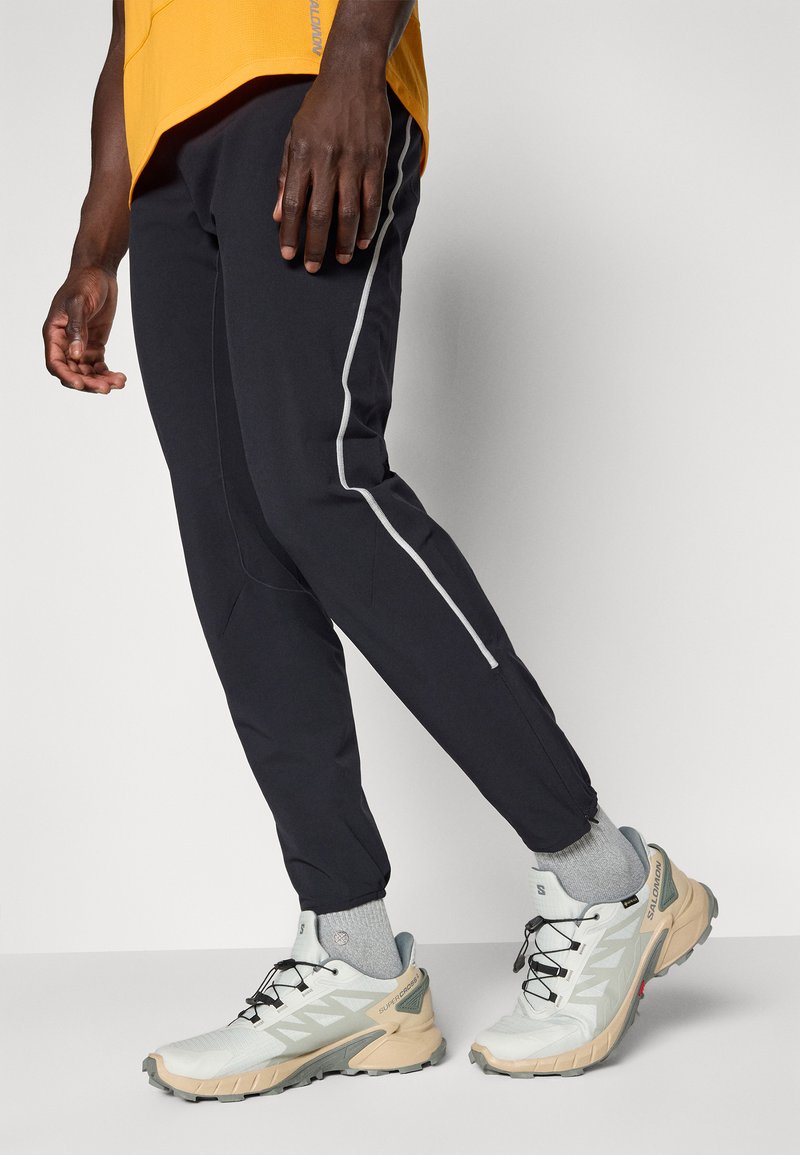 Homme portant un pantalon de sport noir avec une bande latérale blanche, des chaussettes grises et des chaussures de trail Salomon blanches-beiges, se tenant sur un sol gris.