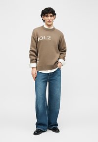 Νέος άνδρας με σγουρά μαύρα μαλλιά, φορώντας ένα καφέ πουλόβερ MOLZ πάνω από λευκό πουκάμισο, παντελόνι τζιν φαρδιάς γραμμής και μαύρα παπούτσια, στέκεται με το ένα χέρι στην τσέπη.