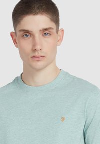 Helle blaue, kurzärmlige T-Shirt aus weichem Stoff, mit einem dezenten orangefarbenen Logo auf der linken Brust. Runder Ausschnitt und lässige Passform.