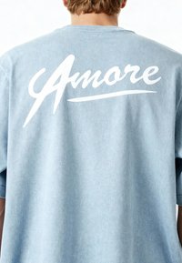 Ljusblå oversized t-shirt med en vit kursiv "Amore" grafik på baksidan. Tillverkad av mjukt, texturerat tyg med korta ärmar.