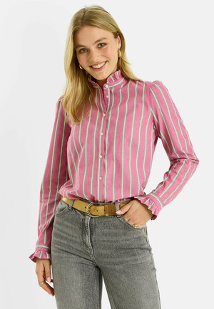 Femme souriante aux longs cheveux blonds portant une chemise rose à rayures vertes avec des poignets à volants, rentrée dans un jean gris avec une ceinture beige.