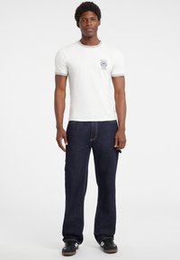Joven de pie, vistiendo una camiseta blanca de manga corta con logotipo, jeans azul oscuro y zapatillas negras, contra un fondo blanco liso.