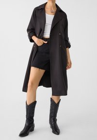 Lang zwarte trenchcoat met knoopaccenten en opgerolde mouwen, gecombineerd met een witte top, zwarte denim shorts en zwarte cowboylaarzen.